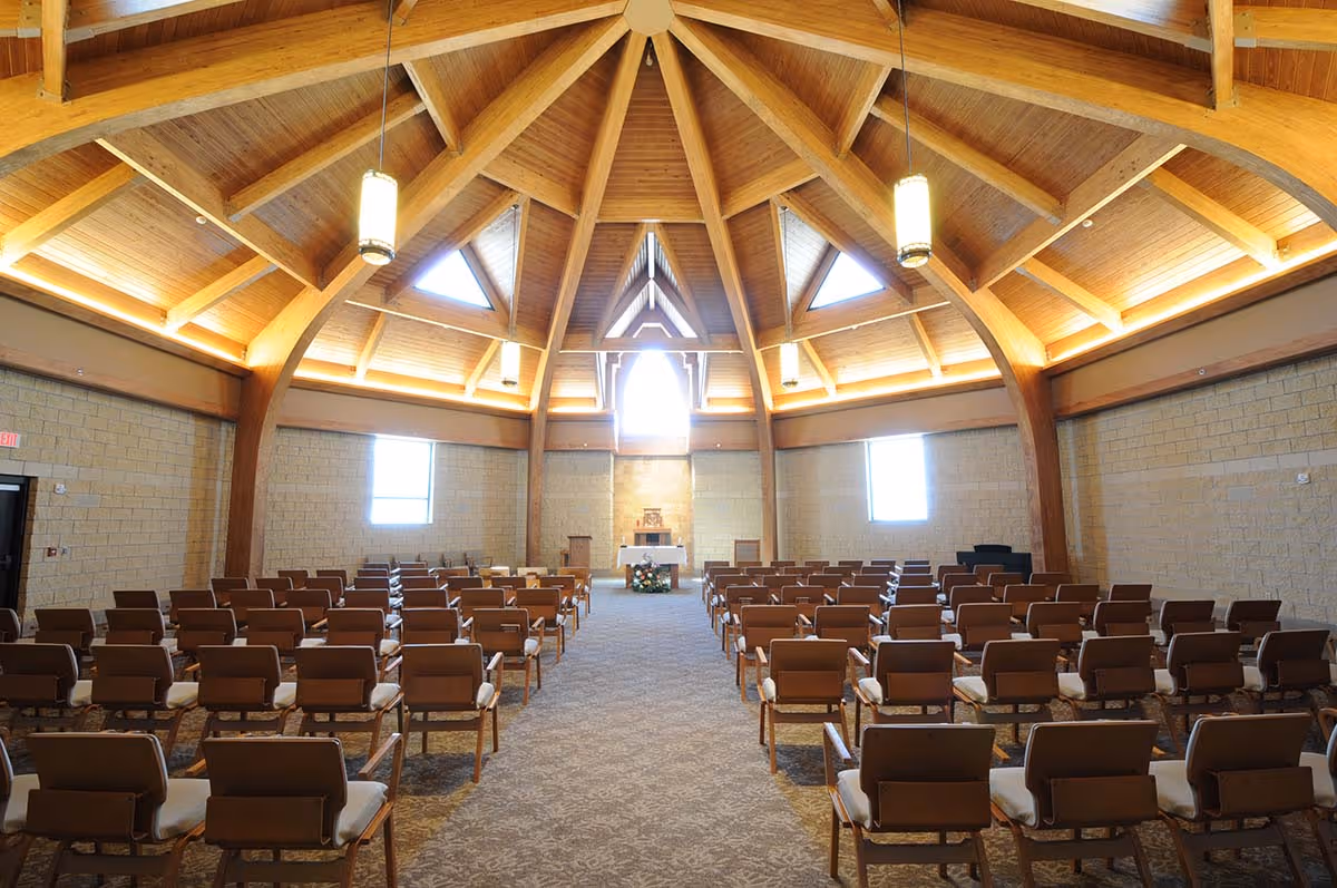 Sunlit chapel-style gathering room with rows of chairs facing an altar under a wooden vaulted ceiling.