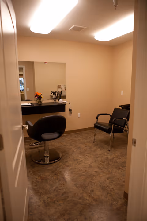 Small salon-style room with a styling chair, wall mirror, counter with tools and a vase of flowers, and two guest chairs in a beige interior.