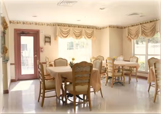 A bright dining room with several tables covered with beige tablecloths and surrounded by wooden chairs with cushioned seats. The room has large windows with decorative valances allowing natural light to fill the space. There is a door with a window on the left side, and the walls are painted in light colors with a floral border near the ceiling.