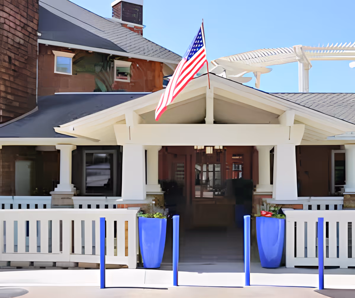 Entrance of a building with a white porch and railing, two tall blue planters with greenery on either side of the entrance, and an American flag mounted above the porch roof. The building has a dark roof and brown siding with windows reflecting the surroundings.
