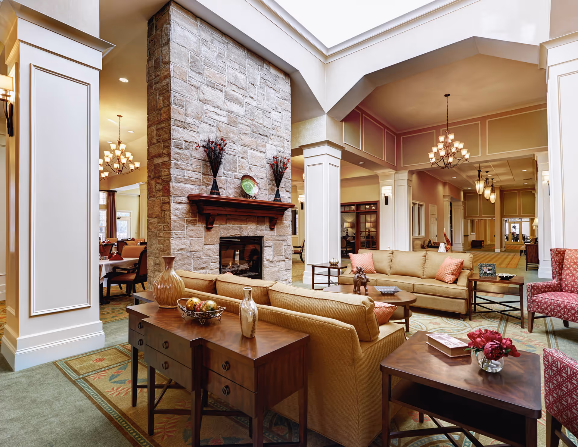 Bright, upscale communal lounge with sofas, armchairs, a central stone fireplace, chandeliers, and decorative tables.