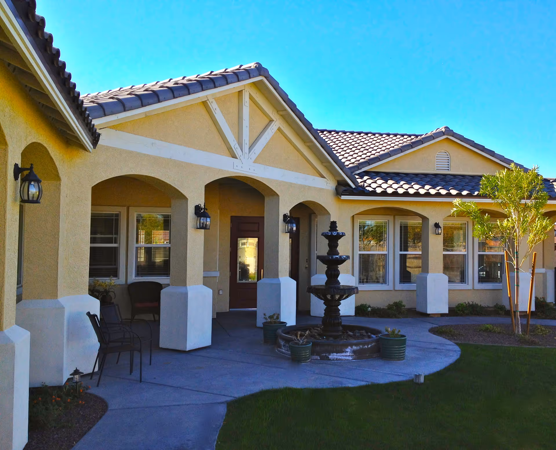 Exterior view of a single-story building with beige stucco walls and a tiled roof under a clear blue sky. The building features arched doorways and windows with white trim, outdoor wall-mounted lantern lights, a small patio area with chairs, a three-tiered black fountain, and some landscaping including a small tree and grass.