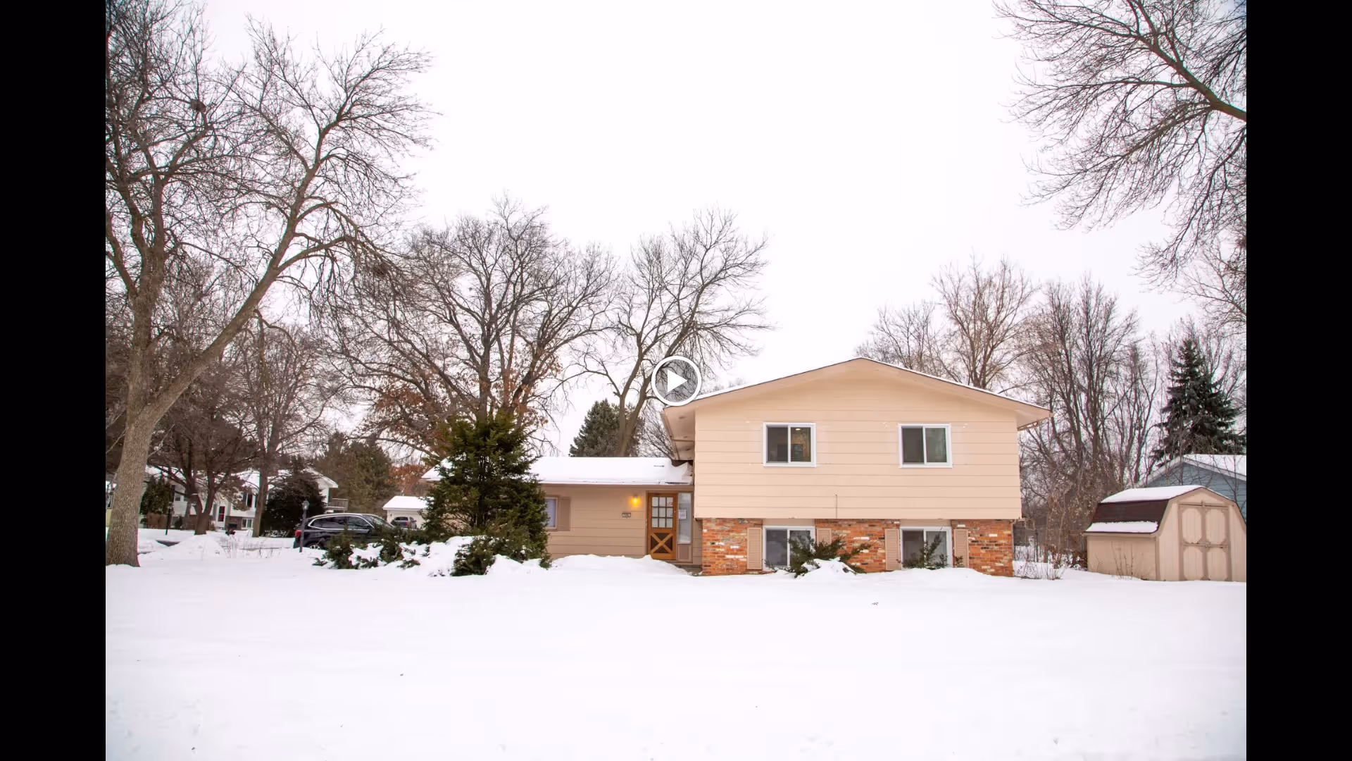 Front view of a split-level house and a small shed in a snowy yard with bare trees.