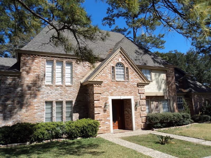 Front exterior view of a two-story brick house with multiple windows, a peaked roof, and a wooden front door. The house is surrounded by green bushes, a lawn, and tall pine trees under a clear blue sky.