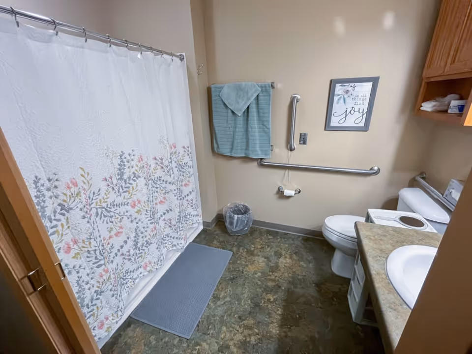 A bathroom with a floral shower curtain, a gray bath mat, a towel rack with two green towels, a toilet with grab bars on the wall, a countertop with a sink, and a framed wall art that says 'In all things find joy'.