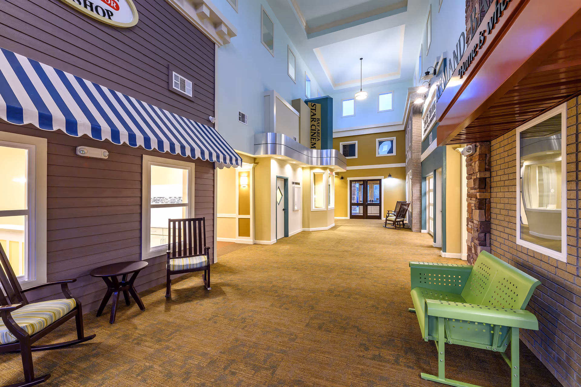 Indoor hallway designed to look like an outdoor street with storefronts, featuring rocking chairs, a green bench, and signs for a barber shop and a cinema. The ceiling is high with windows letting in natural light.