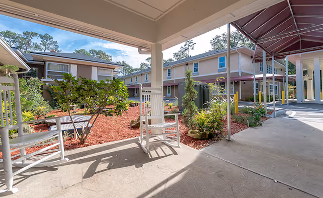 Outdoor covered patio area with white rocking chairs overlooking a landscaped garden with mulch, shrubs, and small trees. Surrounding buildings with beige siding and multiple windows are visible under a blue sky with some clouds.