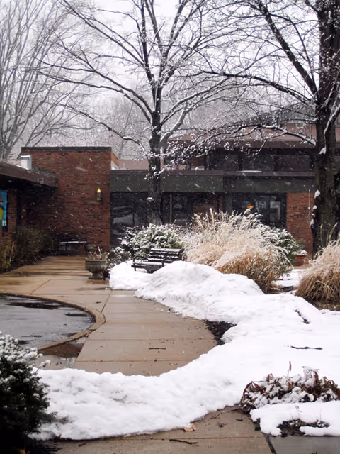 Snow-covered walkway and benches leading to the brick entrance of a single-story building surrounded by bare trees and shrubs.