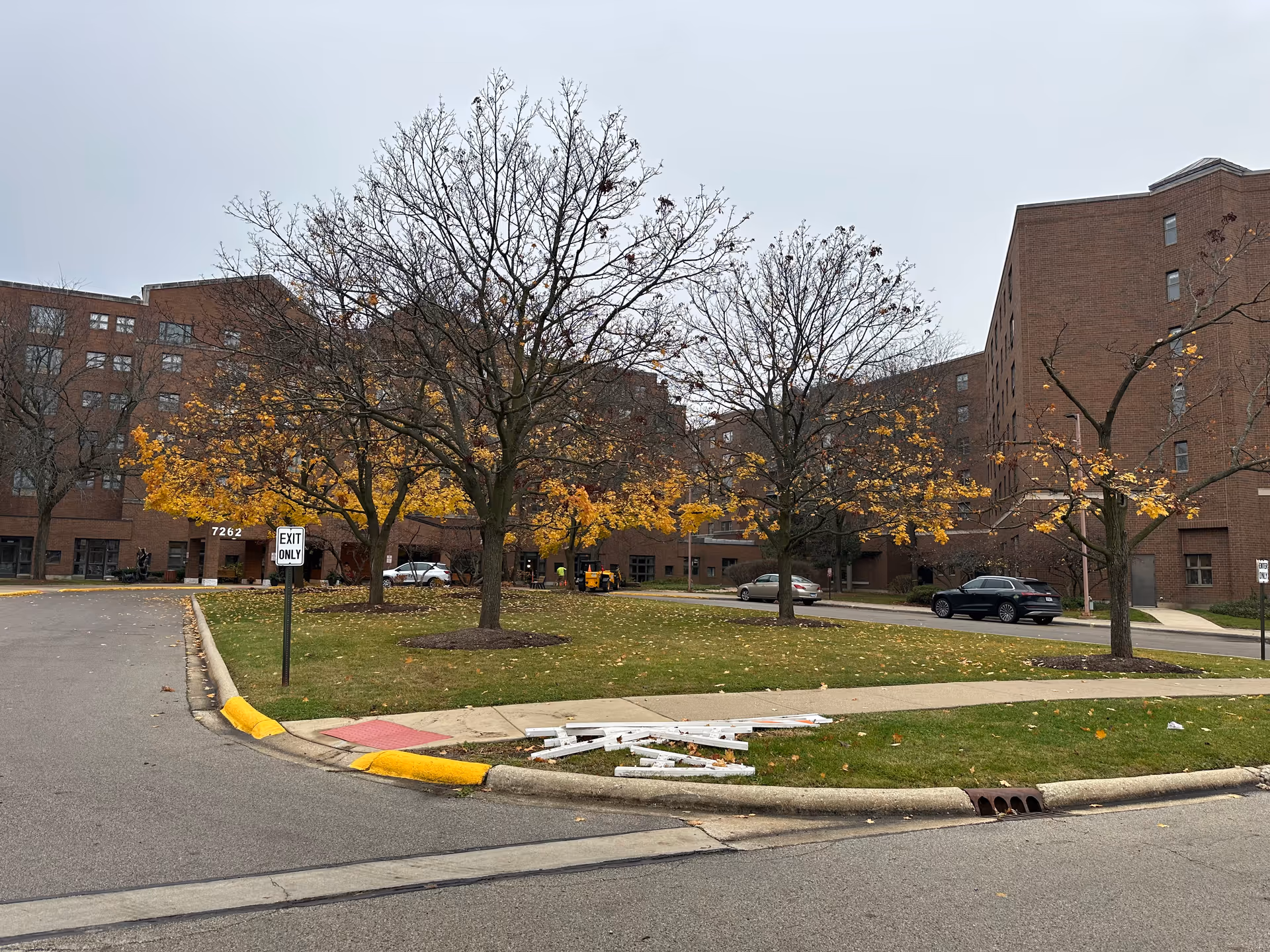 Exterior view of a large brick building with multiple floors, surrounded by leafless trees with some yellow autumn leaves. There is a grassy area with a pile of white wooden debris near the curb. Several cars are parked along the driveway, and a sign reads 'EXIT ONLY'. The sky is overcast.