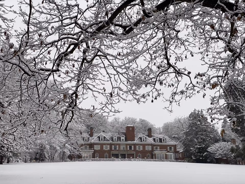 A large brick building with multiple chimneys and dormer windows is seen in the distance, surrounded by snow-covered trees and a wide snow-covered lawn. Snow blankets the branches of the trees in the foreground, creating a wintery scene.