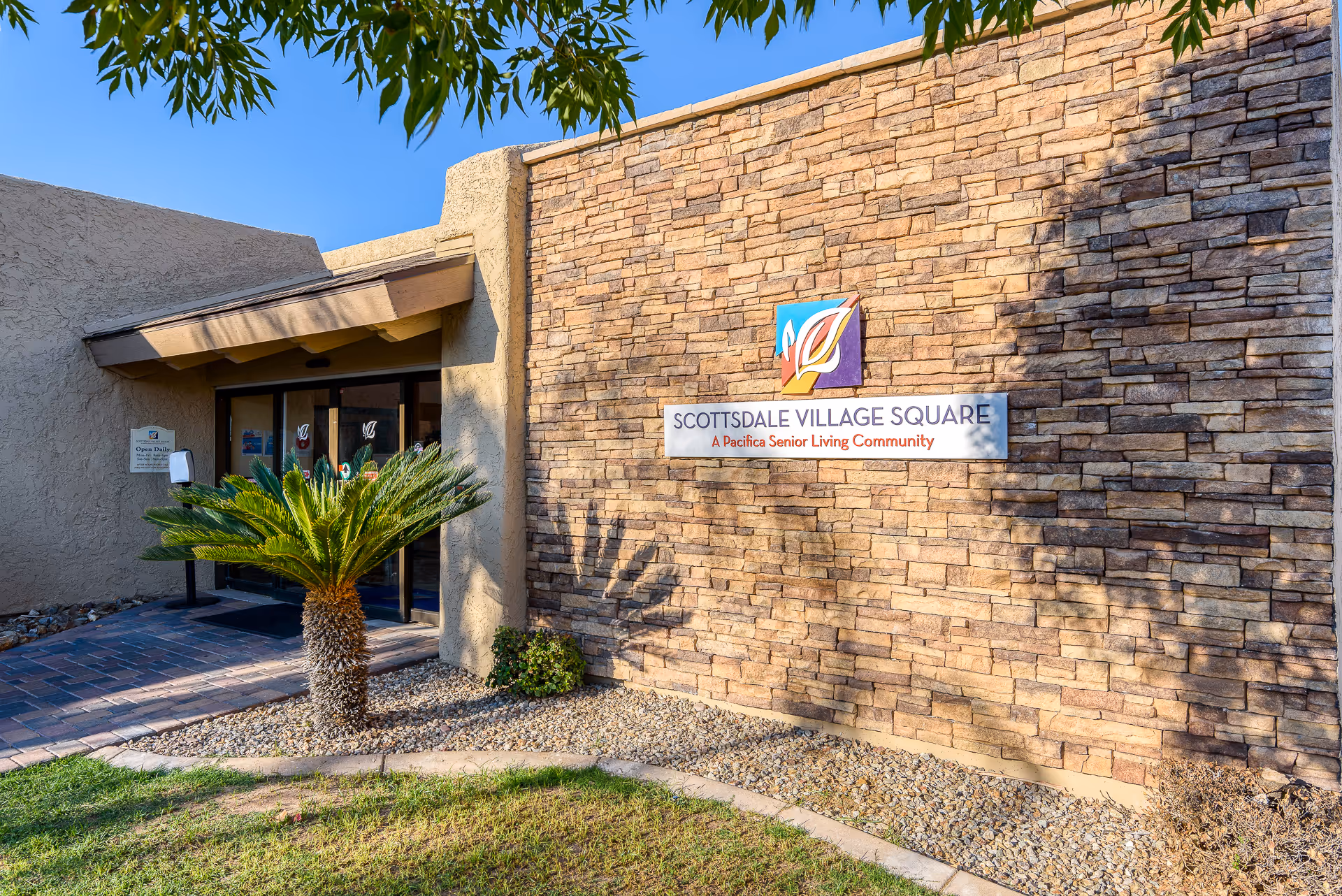Exterior view of Scottsdale Village Square senior living community entrance with a stone wall, a sign displaying the facility name and logo, a small palm tree, and a clear blue sky.