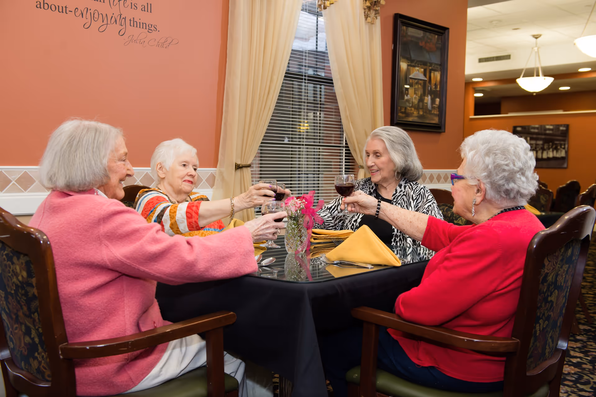 Four elderly women sitting around a dining table in a senior living facility, raising glasses of red wine in a toast. The room has warm-colored walls, a window with curtains, framed artwork, and a table set with yellow napkins and a small floral centerpiece.