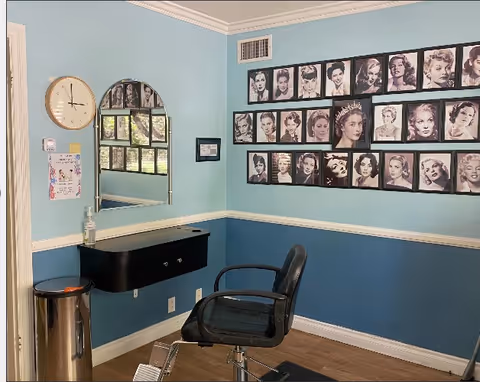 Interior of a small salon or grooming area with a black salon chair in front of a wall-mounted black counter with a mirror above it. The walls are painted light blue on top and darker blue on the bottom with a white chair rail in between. A clock and a hand sanitizer dispenser are mounted on the wall to the left. The wall to the right features a large collage of black and white portraits of various women.