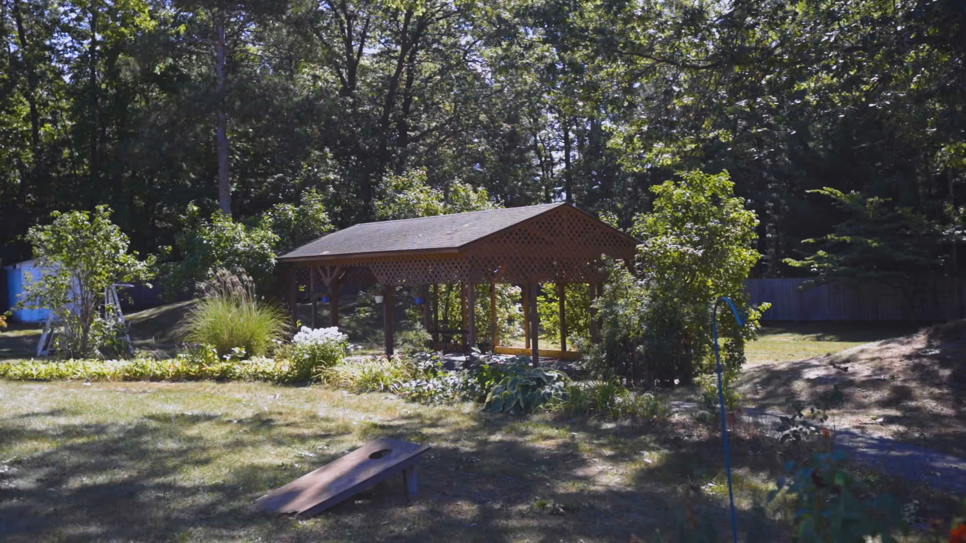 A peaceful outdoor garden area with a wooden gazebo surrounded by various plants and trees. There is a cornhole board set up on the grassy lawn in the foreground, and a wooded area in the background providing shade.