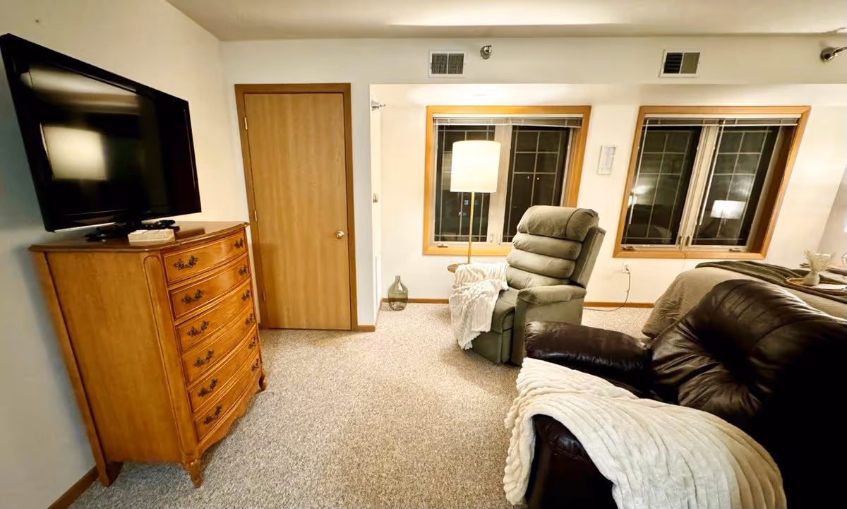 A cozy living space featuring a wooden dresser with a flat-screen TV mounted above it, a green recliner chair with a white throw blanket, a dark brown leather sofa also draped with a white blanket, two windows with wooden frames and blinds, and a floor lamp positioned between the windows.