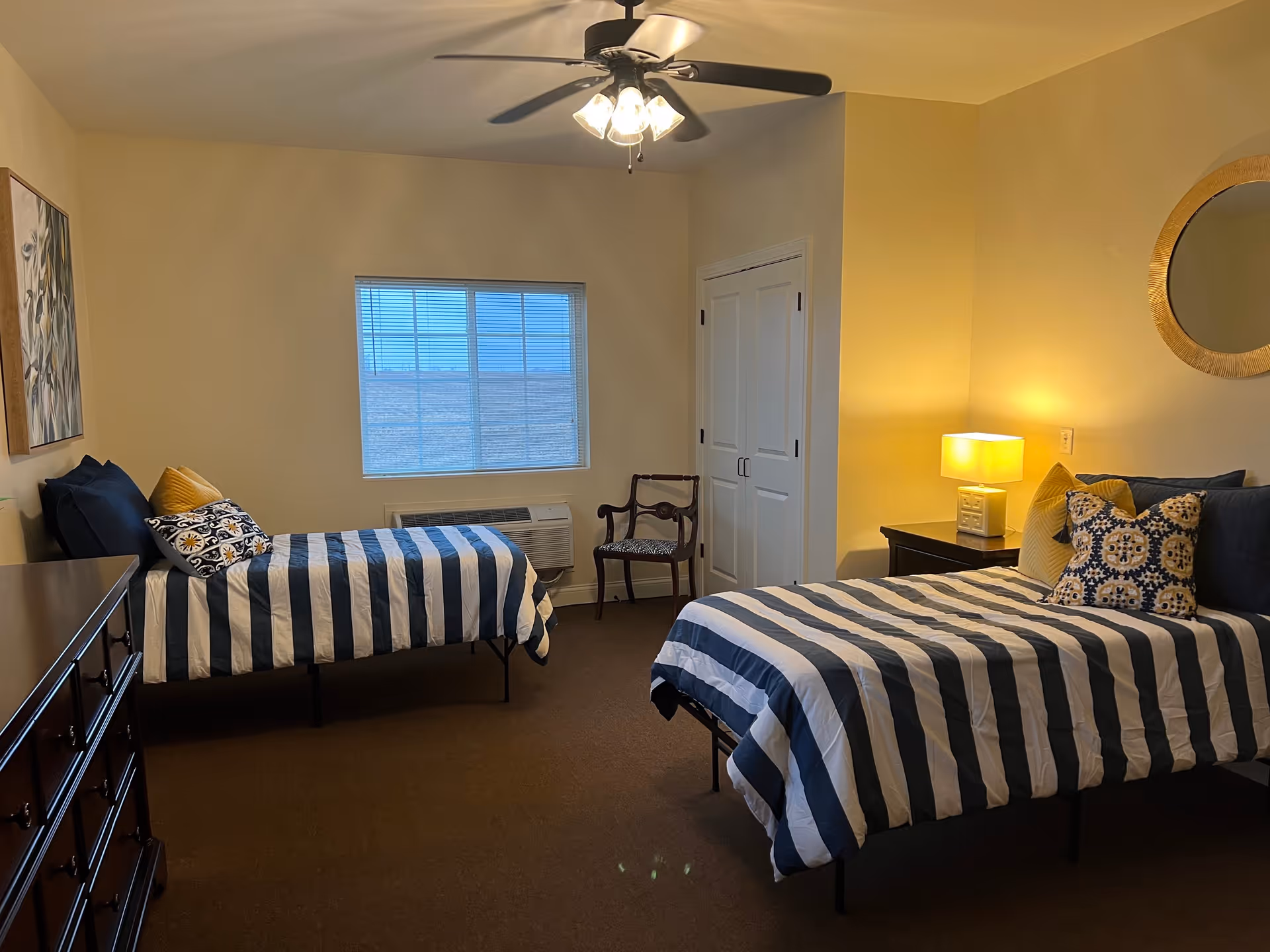 A bedroom with two twin beds featuring navy blue and white striped bedding, decorative pillows, a wooden dresser, a chair, a nightstand with a lamp, a round mirror on the wall, a ceiling fan with lights, and a window with blinds.