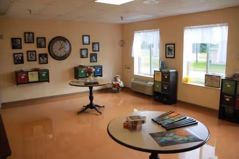 A small activity room in a senior facility with round tables, wall art, and windows.