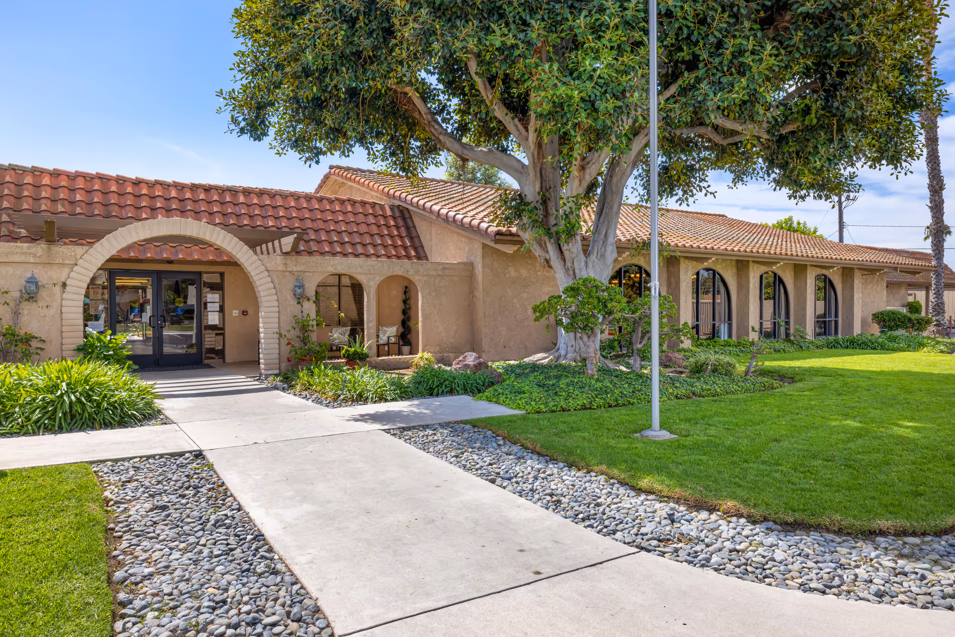 Exterior view of Brookdale Garden Grove facility featuring a beige stucco building with a red tile roof, arched entryway, large windows, well-maintained green lawn, landscaping with bushes and a large tree, and a concrete walkway leading to the entrance.