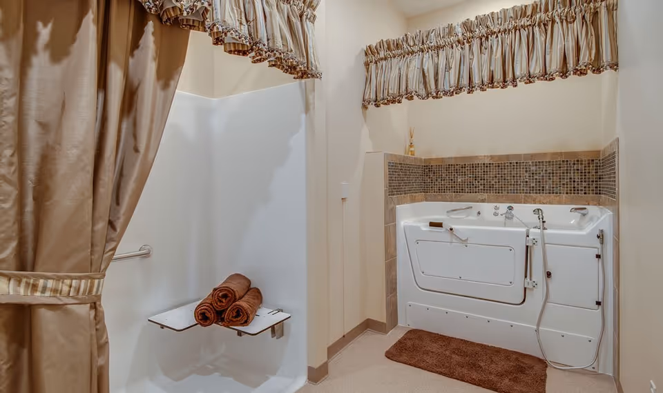 A senior living facility bathroom featuring a walk-in shower with a fold-down seat and three rolled brown towels placed on it. Next to the shower is a walk-in bathtub with safety handles and a handheld showerhead. The walls are light-colored with a decorative tile border above the bathtub. Brown curtains and a matching brown bath mat complete the setup.