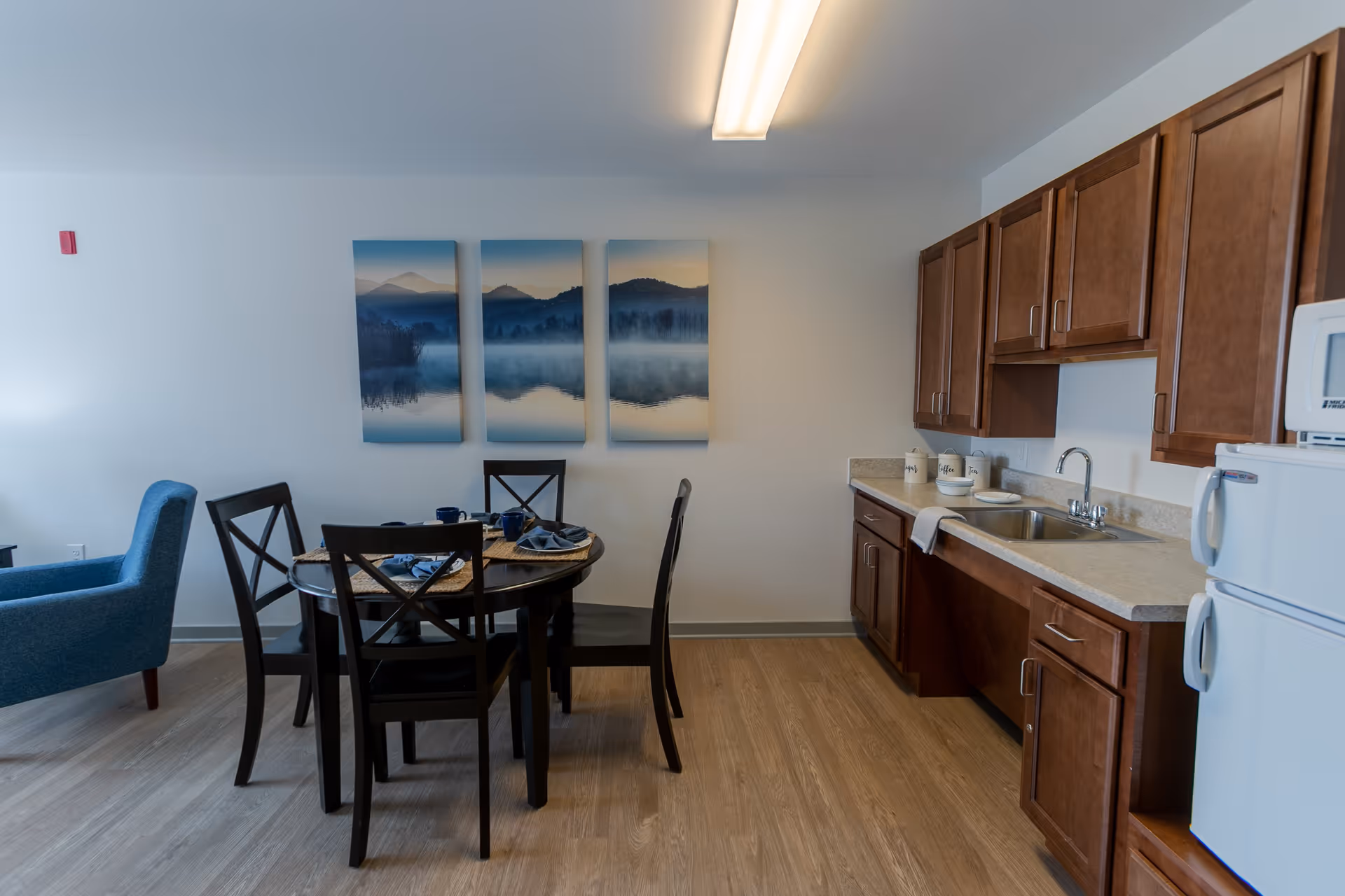 Interior view of a senior living facility kitchen and dining area at Oasis at 56th. The room features wooden cabinets, a countertop with a sink, a white refrigerator, and a microwave. A round dining table with four black chairs is set with placemats, napkins, and cups. A blue armchair is visible to the left, and a three-panel landscape artwork hangs on the wall above the dining table.