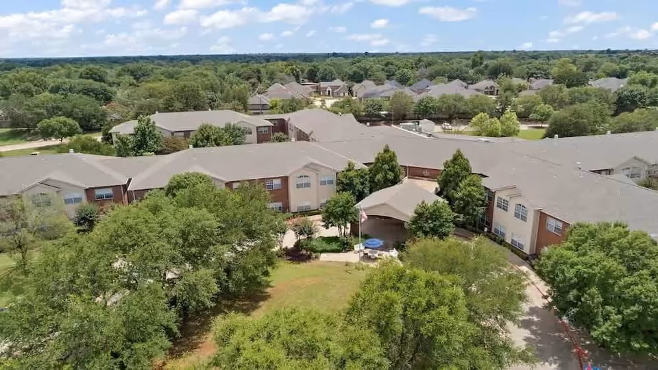 Aerial view of Morada Lake Arlington senior living facility showing multiple connected buildings surrounded by trees and greenery under a partly cloudy sky.