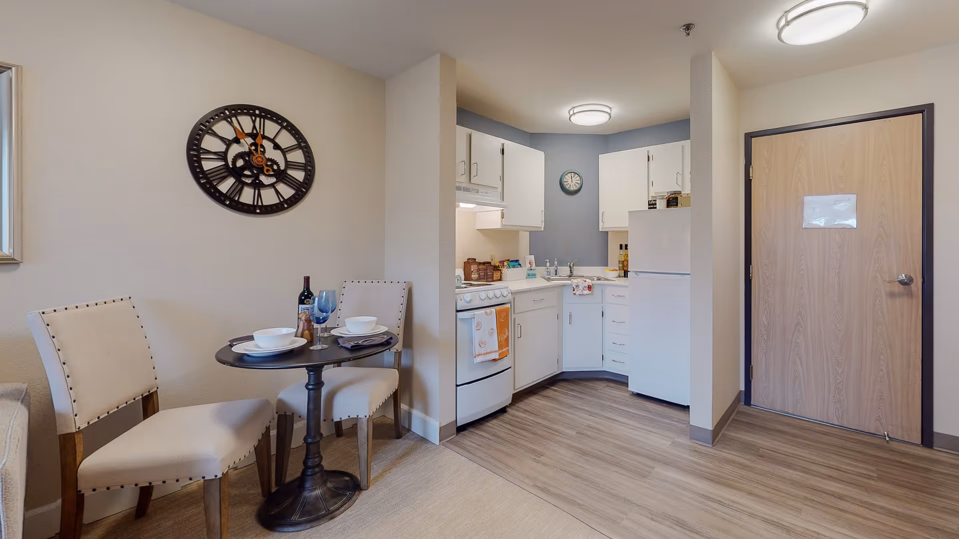 Small dining area with a round table set for two with white bowls, plates, and a wine bottle, next to a compact kitchen featuring white cabinets, a stove, refrigerator, and a wooden door in the background.