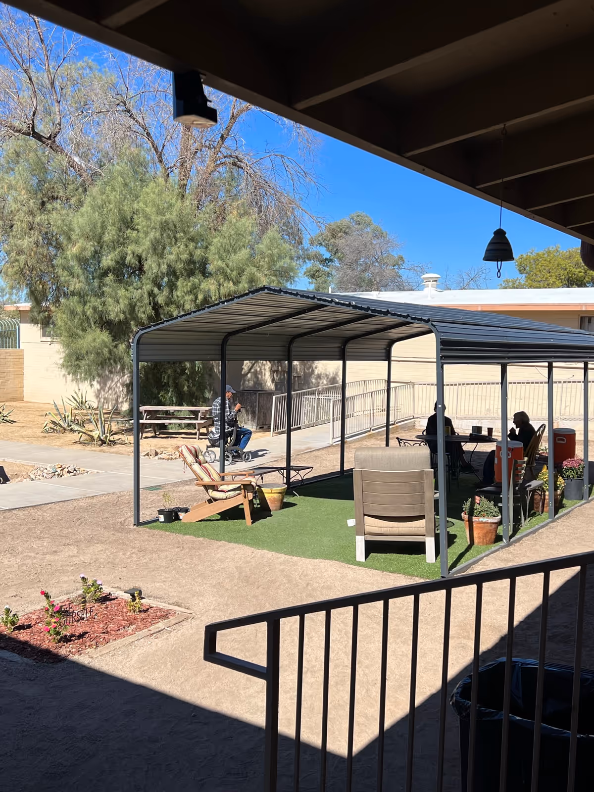 Outdoor covered seating area with chairs and a table on artificial grass, surrounded by potted plants. Two people are sitting at the table under the metal canopy, and another person is riding a mobility scooter nearby. Trees and a building wall are visible in the background under a clear blue sky.
