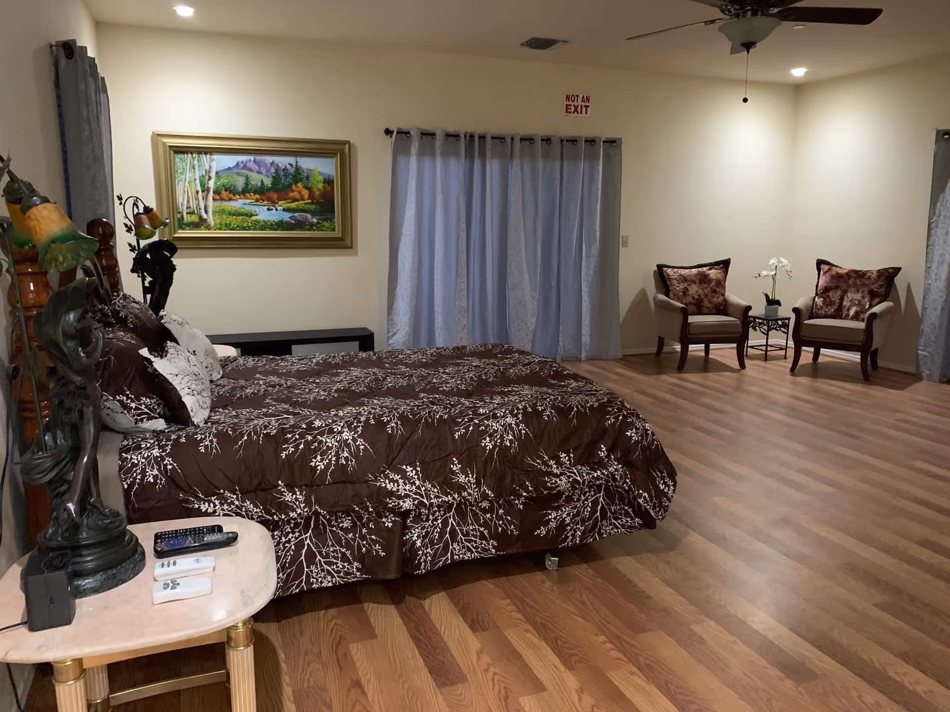 A spacious bedroom with a large bed covered in a brown and white patterned comforter. There is a small table beside the bed with remote controls and a decorative lamp. On the far wall, there is a framed landscape painting above a black bench. Two armchairs with floral cushions and a small table with a white orchid are placed in a corner. The room has wooden flooring, light-colored walls, and curtains covering a window or door. A ceiling fan with a light is visible on the ceiling.