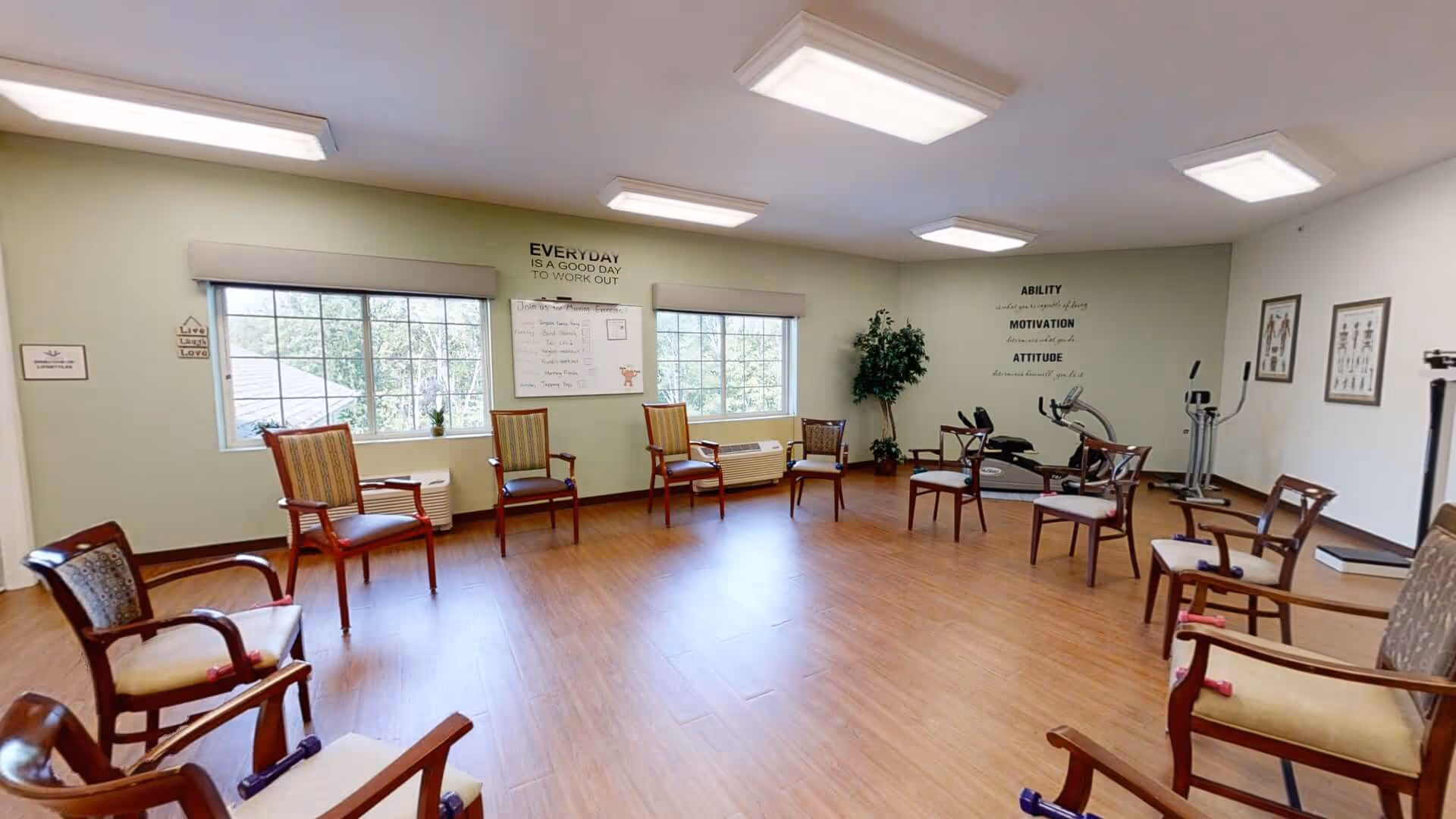 A bright activity/exercise room with chairs arranged around the perimeter, large windows, and a few pieces of exercise equipment against the far wall.
