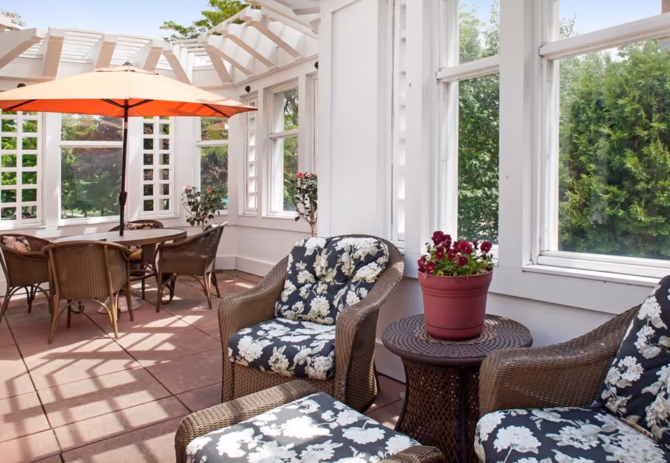 Enclosed sunroom with wicker seating and floral cushions, a small table with a potted plant, and a dining table under an orange umbrella.