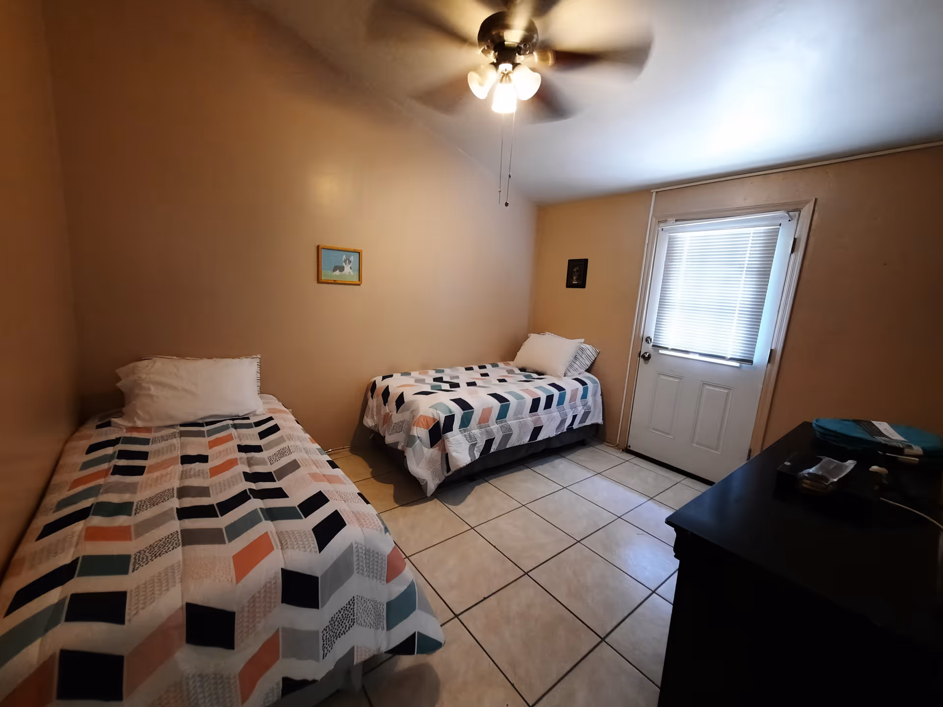 A small bedroom with two single beds covered in colorful geometric patterned bedspreads. There is a ceiling fan with lights above, a door with a window covered by blinds, and a black dresser with some items on top. The walls are painted beige and the floor is tiled.
