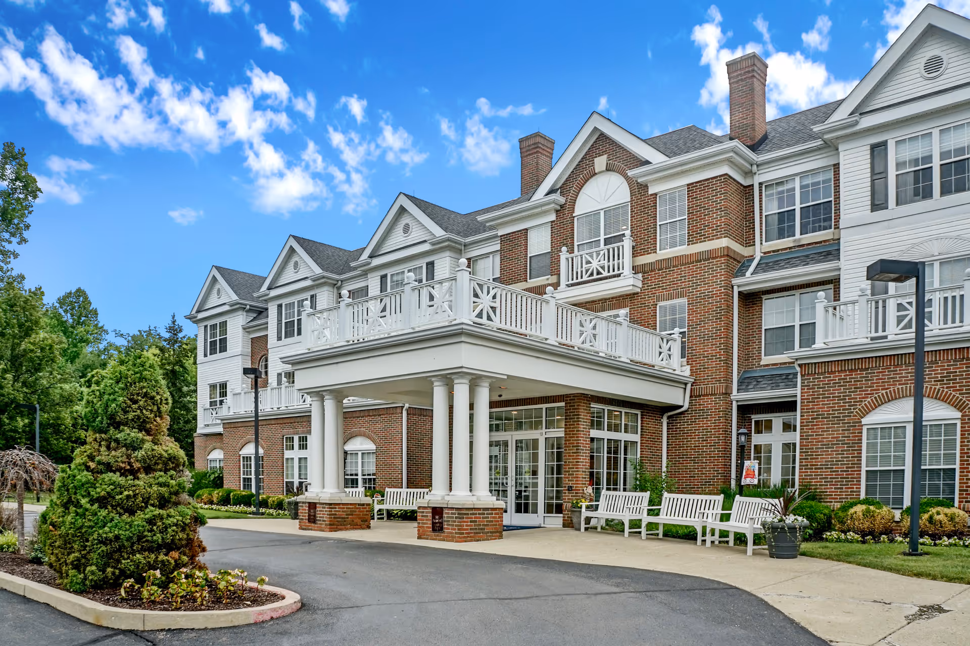 Exterior view of a senior living facility with a brick and white facade, featuring a covered entrance supported by columns, multiple windows, white benches outside, landscaped greenery, and a clear blue sky with scattered clouds.