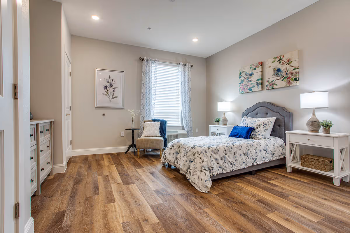 A bright and cozy bedroom with a single bed featuring floral bedding and a blue accent pillow. The room has wooden flooring, two white nightstands with lamps and small plants, a cushioned chair with a pillow and a blue throw, a window with patterned curtains, and floral artwork on the walls.