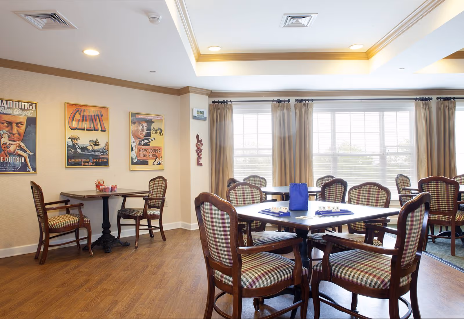 Bright common dining/activity room with wooden tables and upholstered chairs, vintage posters on the wall, and large windows with curtains.