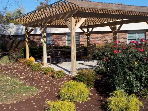 Outdoor seating under a wooden pergola beside a brick building and blooming shrubs.