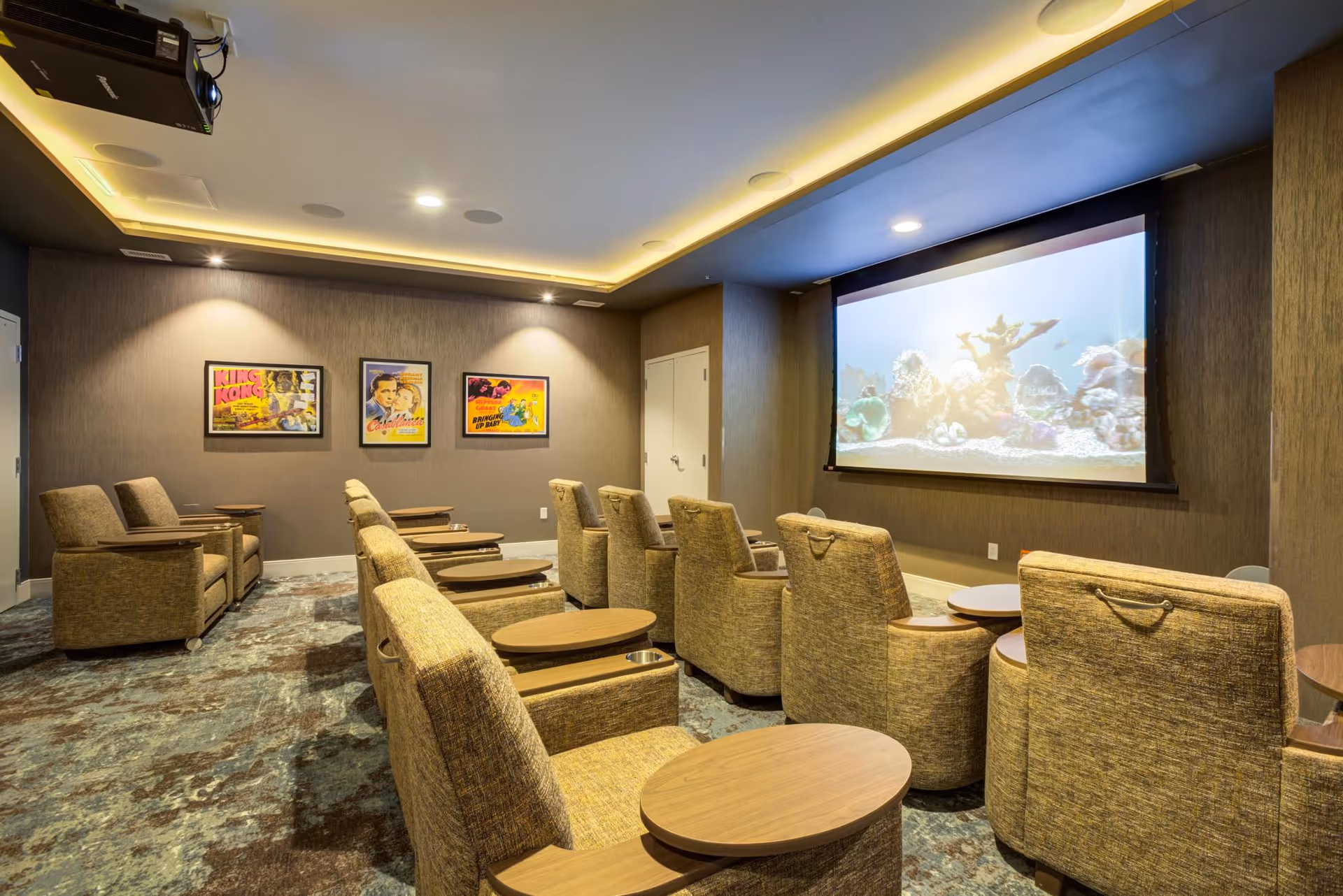 A cozy movie theater room with multiple rows of comfortable beige armchairs, each equipped with a small wooden table. The room has a large projection screen displaying an underwater scene with coral and fish. The walls are decorated with three framed vintage movie posters, and the ceiling features recessed lighting.