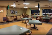 A common area in a senior living facility with several tables covered with green tablecloths and chairs around them. The room has a wooden floor, beige walls, and windows along one side. There are a few armchairs and framed pictures on the wall, with some decorations hanging from the ceiling.