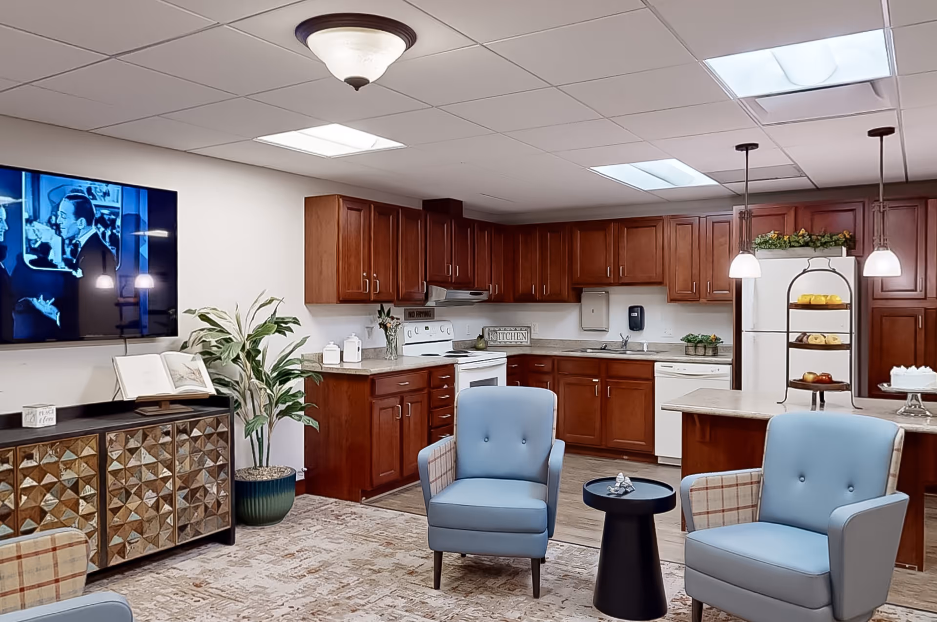 Cozy common room featuring blue armchairs, a wall-mounted TV, and a wood-finished kitchenette with an island and pendant lights.