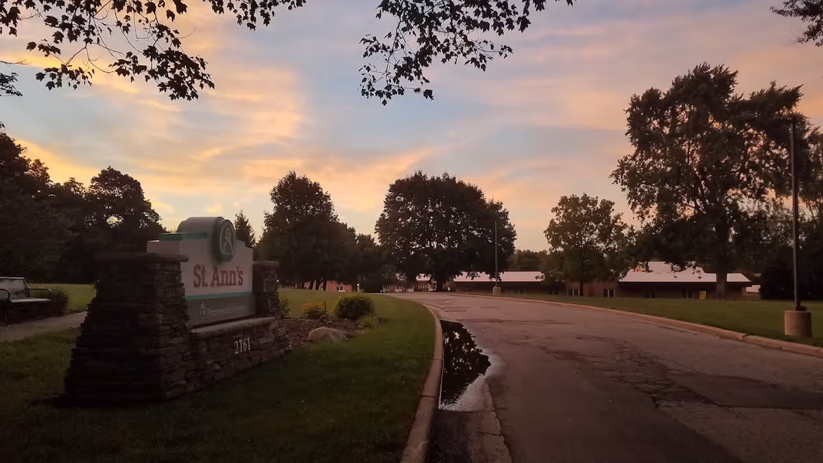 View of the entrance road to St. Ann's Home at sunset, with a stone sign displaying the facility's name and address. Trees and a building with a white roof are visible along the road under a colorful sky.