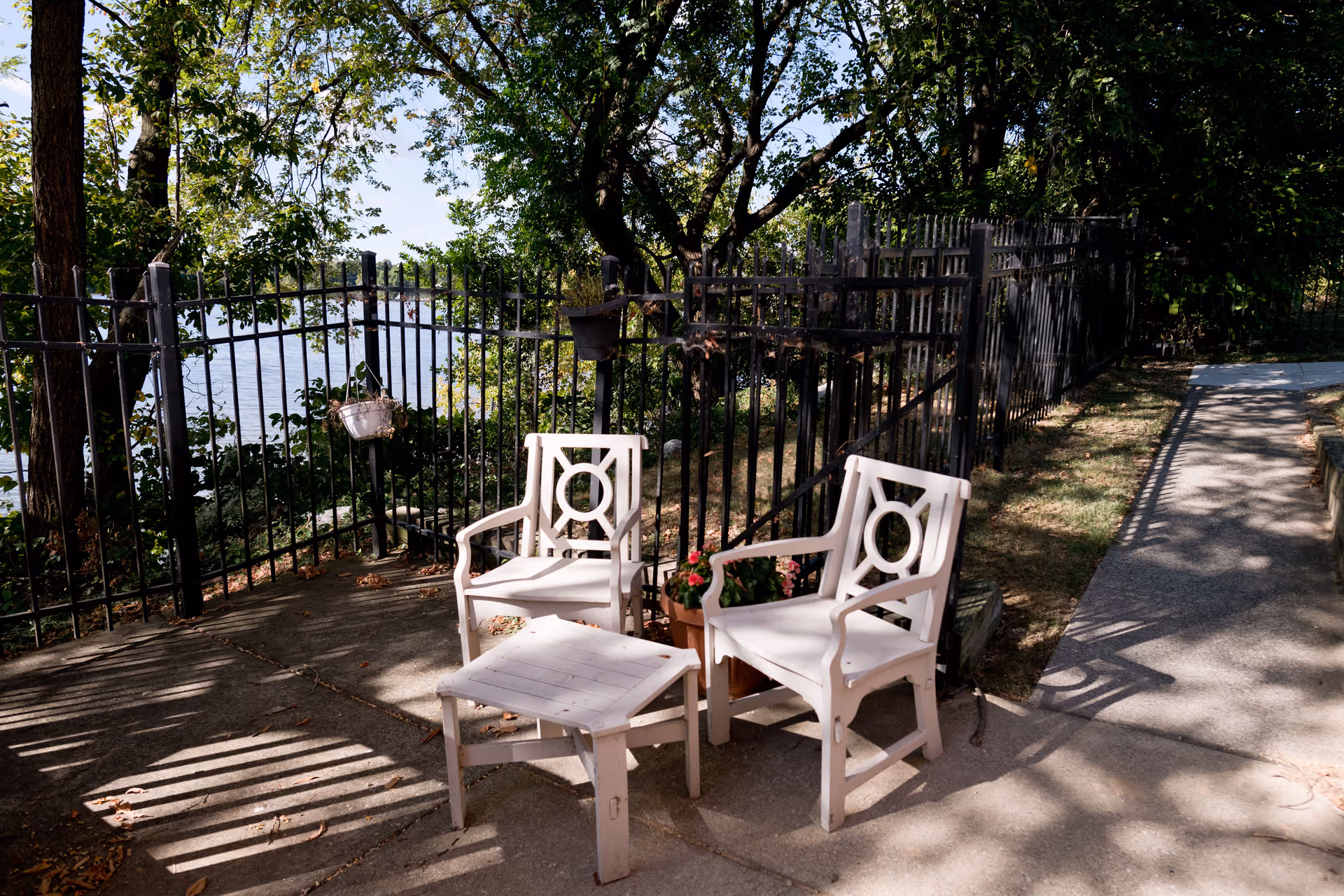 Two white wooden chairs and a small matching table on a concrete patio area next to a black metal fence with trees and greenery in the background, overlooking a body of water.