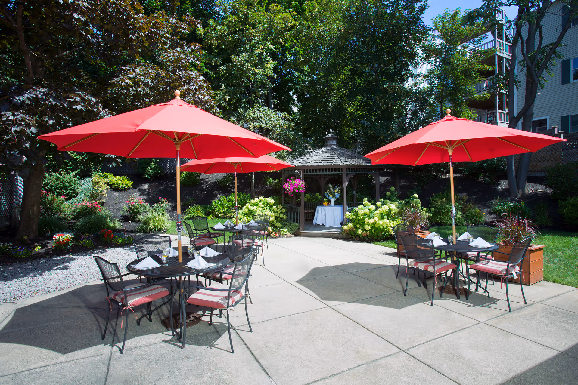 Patio courtyard with round tables and chairs shaded by red umbrellas, a gazebo, and surrounding garden greenery.