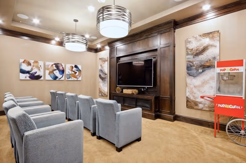 A cozy media room with two rows of gray upholstered chairs facing a large flat-screen TV mounted on a dark wooden wall unit. The room features beige walls with abstract artwork and two modern circular ceiling lights. A red popcorn machine is positioned on the right side of the room.