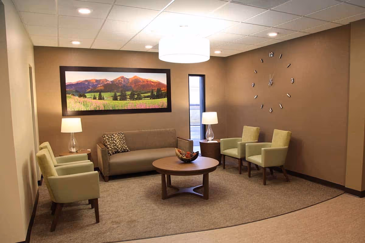 Interior lounge at a senior living facility with a gray sofa, light-green chairs, a round coffee table, lamps, a landscape painting, and a large wall clock.