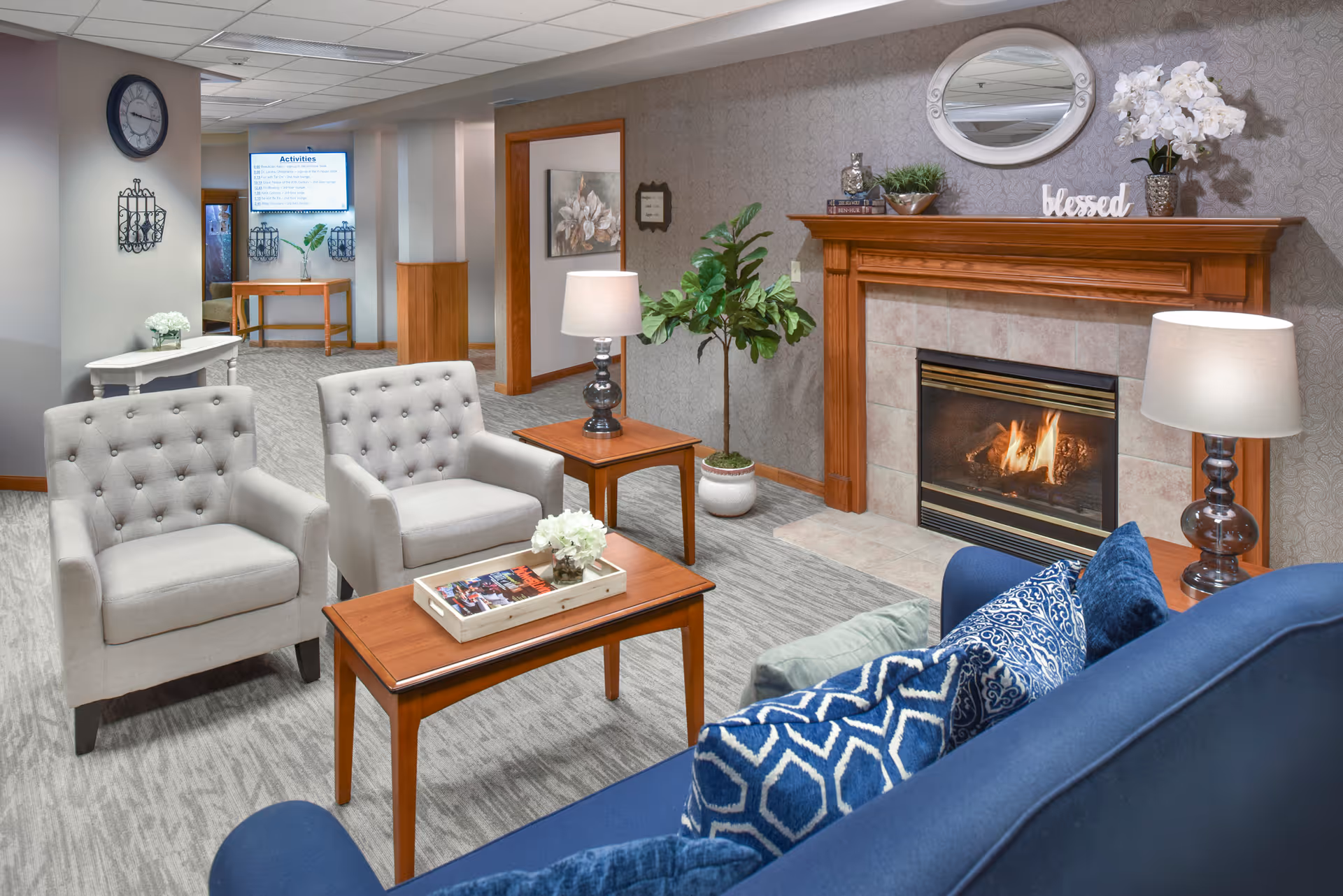 A cozy living room area in a senior living facility featuring a lit fireplace with a wooden mantel decorated with a mirror, flowers, and a 'blessed' sign. The room has a blue couch with patterned pillows, two light gray armchairs, wooden coffee and side tables with lamps and flower arrangements. The background shows a hallway with a clock on the wall and an activities board.