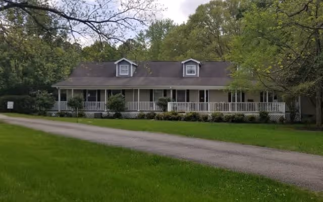 A single-story building with a long porch and white railing, surrounded by green grass and trees under a partly cloudy sky.