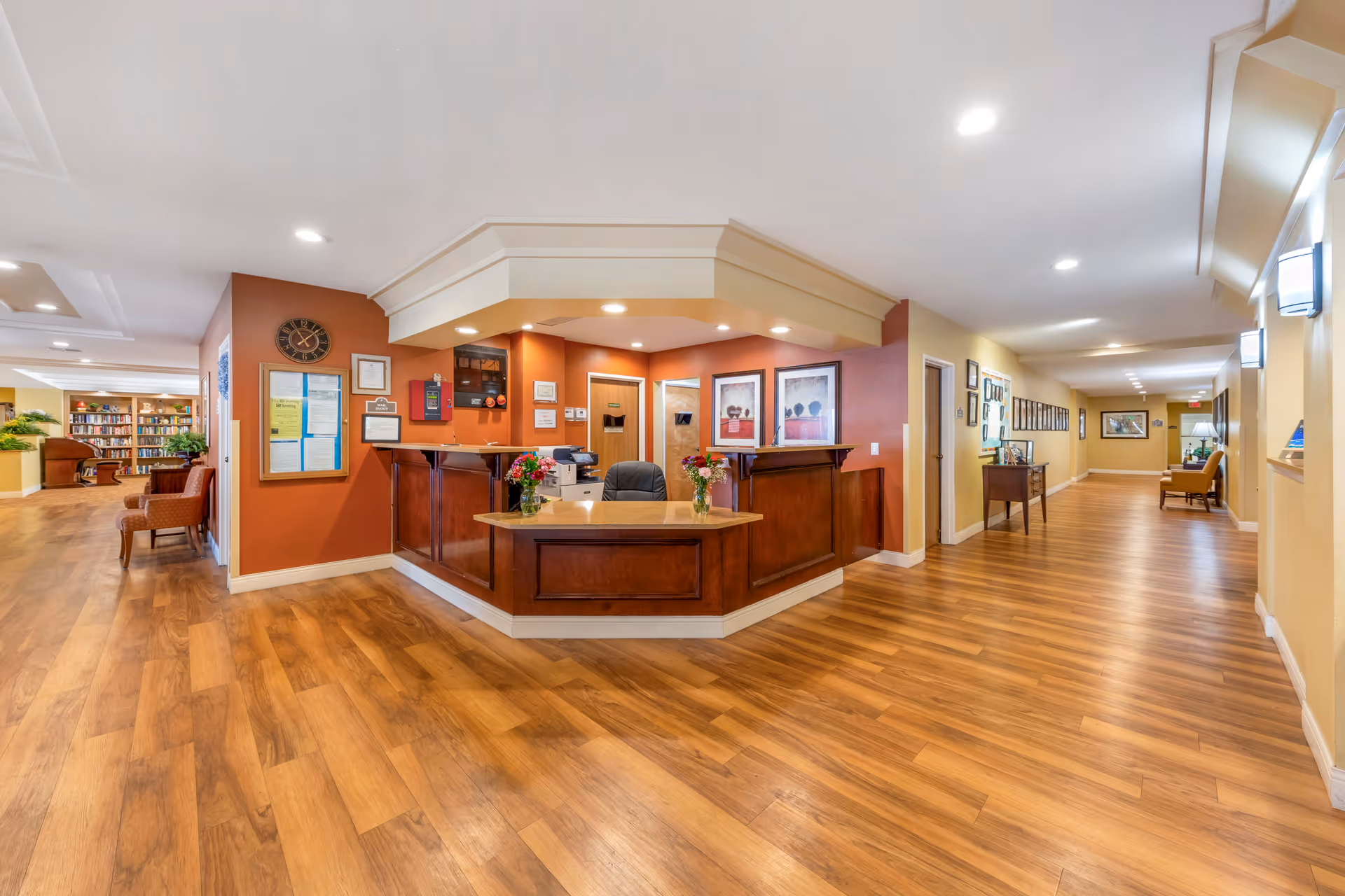 Bright reception desk and lobby with wood floors, seating, framed artwork, and a long hallway.