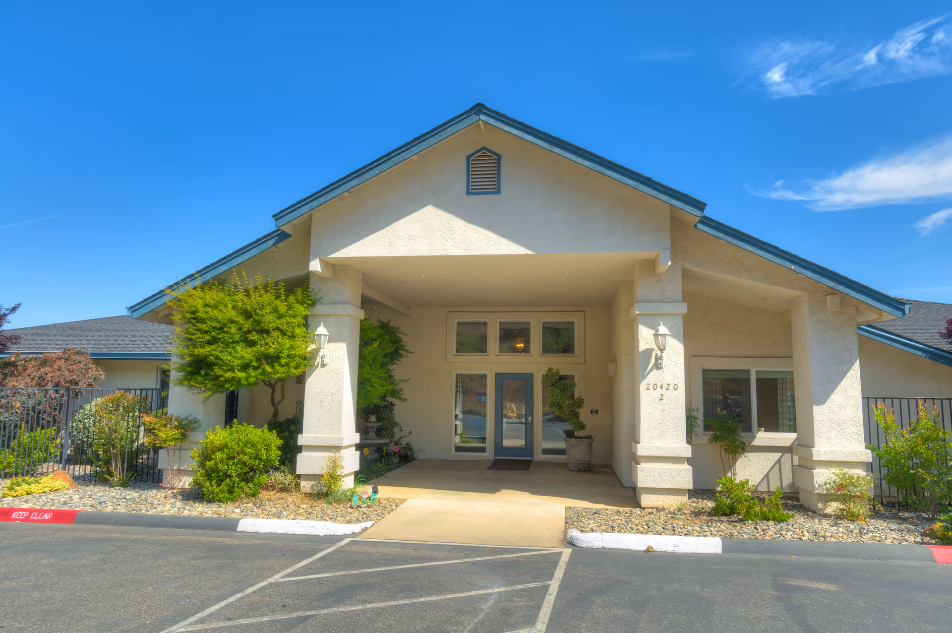 Front exterior view of a single-story building with a covered entrance supported by two columns. The building has beige stucco walls, a blue-gray roof, and several windows. There are small landscaped areas with bushes and trees on either side of the entrance. The sky is clear and blue.