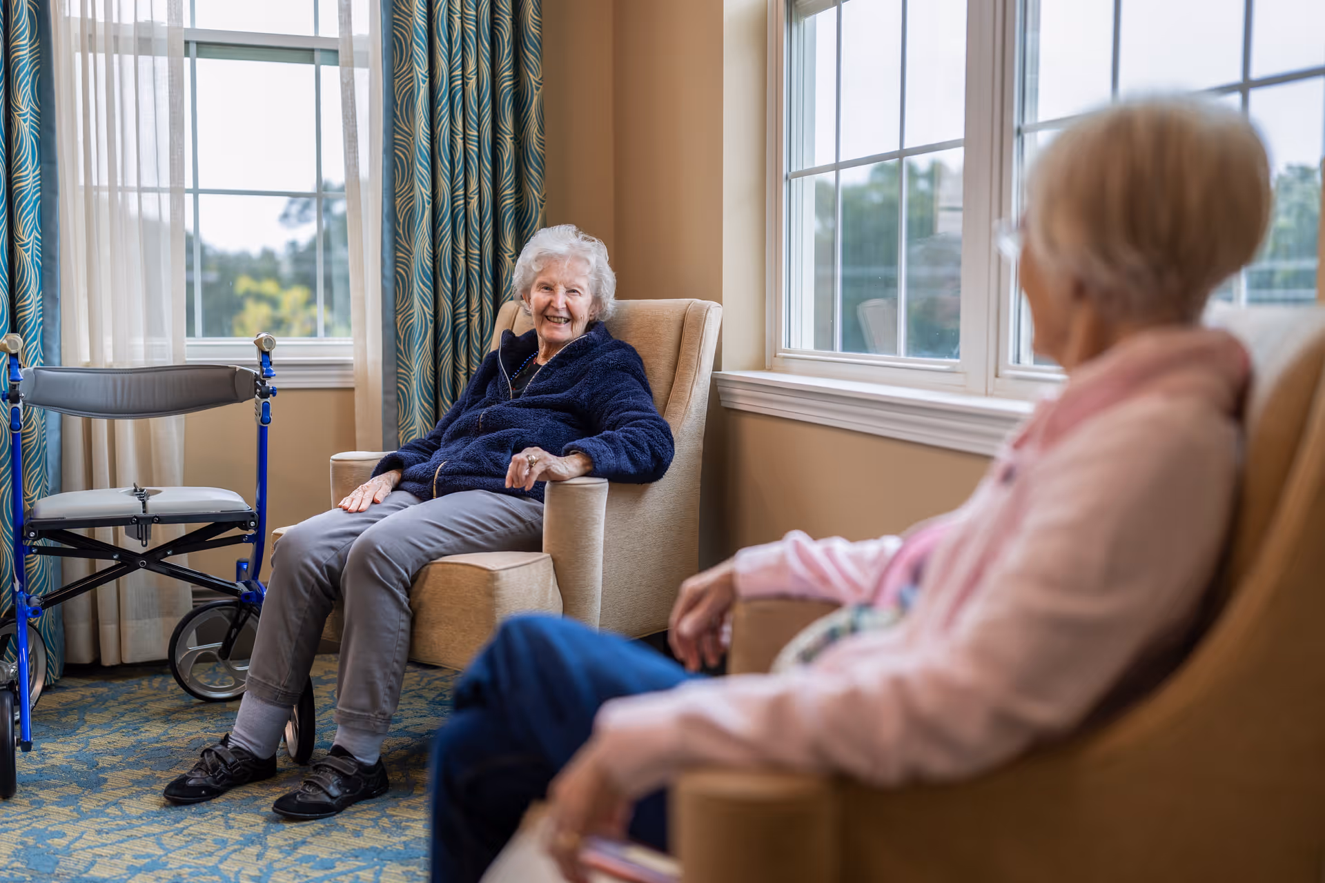 Two elderly women sitting in armchairs in a well-lit room with large windows and patterned curtains. One woman is smiling and wearing a dark blue jacket, while the other woman, wearing a light pink jacket, is seen from behind. A blue walker is positioned near the window.