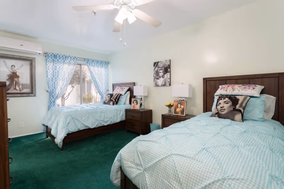 A bedroom with two twin beds featuring light blue checkered bedding and decorative pillows with black and white portraits. Between the beds is a wooden nightstand with two lamps and framed photos. A window with blue patterned curtains lets in natural light, and a ceiling fan is visible above. The room has green carpet and light-colored walls adorned with black and white portrait artwork.