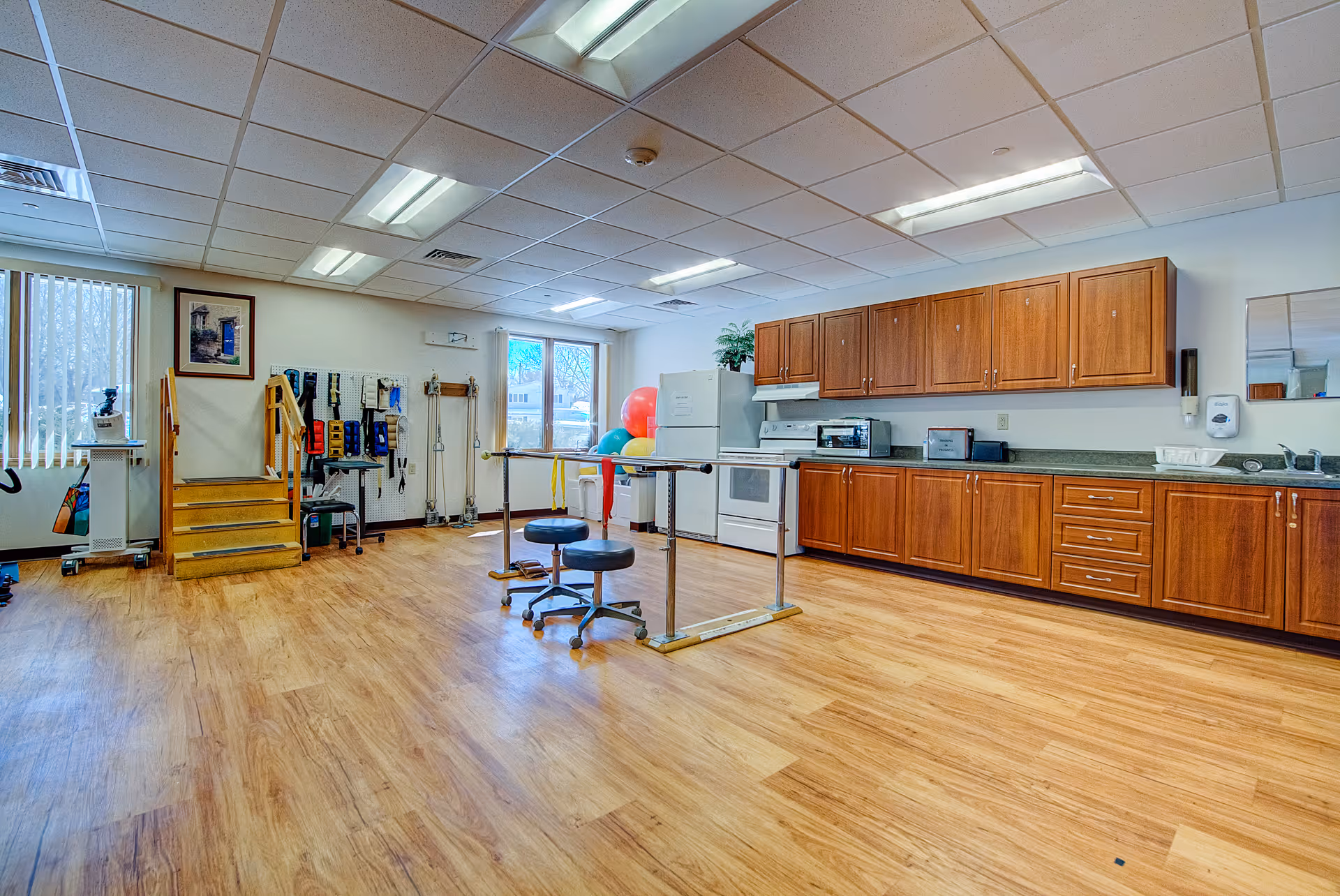 A spacious room with wooden flooring and a drop ceiling with fluorescent lights. On the right side, there is a long countertop with wooden cabinets above and below, a white stove, refrigerator, microwave, toaster, and a sink. In the center of the room, there are two black stools on wheels and parallel bars, likely for physical therapy or rehabilitation exercises. On the left side, there are exercise equipment, therapy tools hanging on the wall, a small set of wooden stairs, and large windows with vertical blinds letting in natural light.